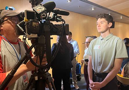 Nolan Cottingham wearing a light green shirt and black pants being interviewed on camera by a news crew. He stands with hands clasped while a cameraman in a red and gray shirt films him. Other people are visible in the background in a brightly lit room with wood-paneled walls.