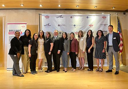 A group of thirteen professionally dressed individuals pose for a group photo on a wooden stage. Behind them is a backdrop featuring logos, including TSMC Arizona, Maricopa Community Colleges, and Northern Arizona University. To the left of the group is a standing banner that reads "Brilliant Together," and to the right is an American flag. The setting appears to be a formal event or ceremony, and the atmosphere is positive and celebratory.