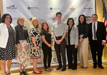 Group of eight people standing and smiling in front of a step-and-repeat banner featuring multiple company logos, including TSMC and Arizona@Work. The group includes both men and women dressed in business or business-casual attire. An American flag is visible on the right side of the image.