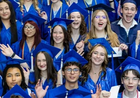 A large, energetic group of dual enrollment students from Rio Salado College cheer and smile during commencement. They are dressed in bright blue graduation gowns and caps, some holding diplomas and raising their hands in celebration. The diverse group of young graduates stands close together, radiating excitement and pride as they mark this academic milestone.